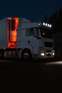 A white truck lit by headlights and interior lights on a dark road in mountainous terrain.